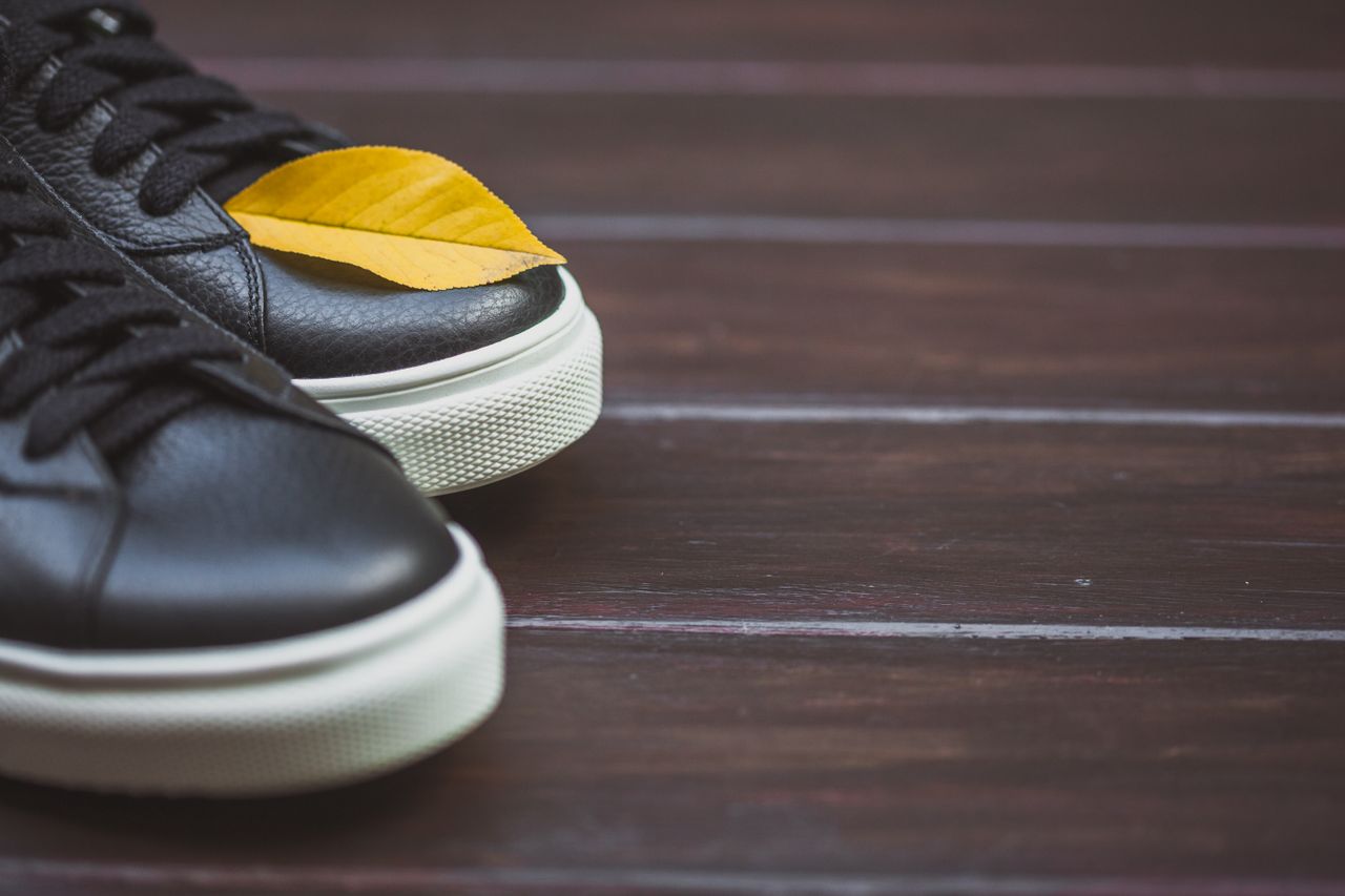 Close-up of black sneakers with a yellow autumn leaf on a wooden floor, symbolizing fall fashion.
